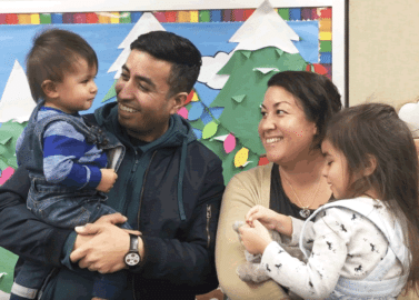 A man and woman smile while holding a young boy and girl in front of a colorful winter-themed bulletin board with snow-capped trees and paper holiday lights.