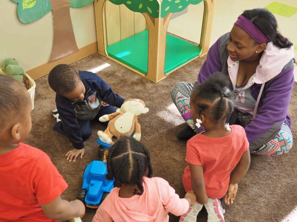 A woman sits on the floor with four young children playing together. Toys, including a stuffed animal and a toy truck, are on the carpet. The group is indoors, near a wooden play area decorated with a tree motif.