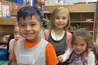 Four young children wearing backpacks smile at the camera in a classroom setting with shelves and educational materials in the background.