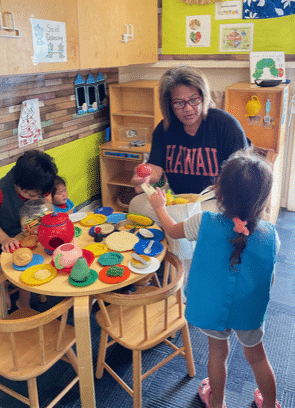 A woman and three young children play with colorful toy food at a small table in a classroom decorated with posters and art. The woman engages with the children as they pretend to share a meal together.