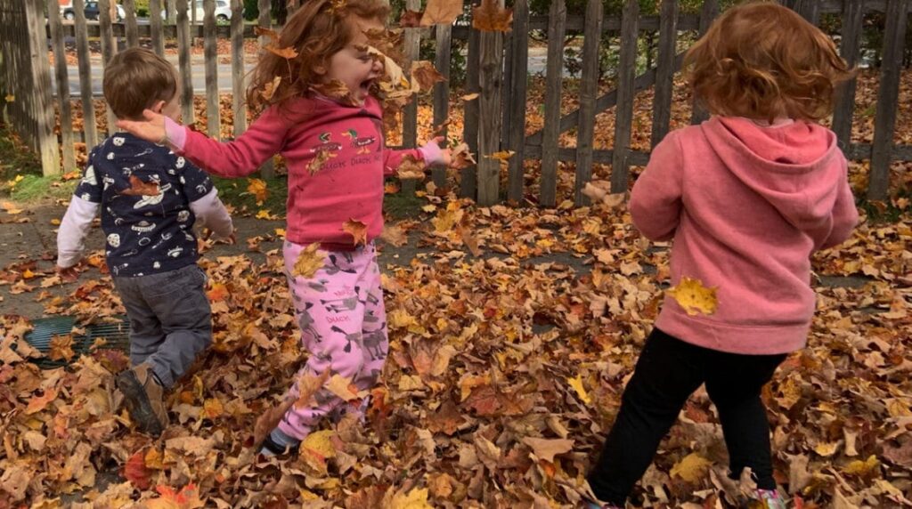 Three young children play joyfully in a pile of fallen autumn leaves, with two girls in pink and a boy in a dark shirt. Leaves are scattered in the air and on the ground, and a wooden fence is in the background.