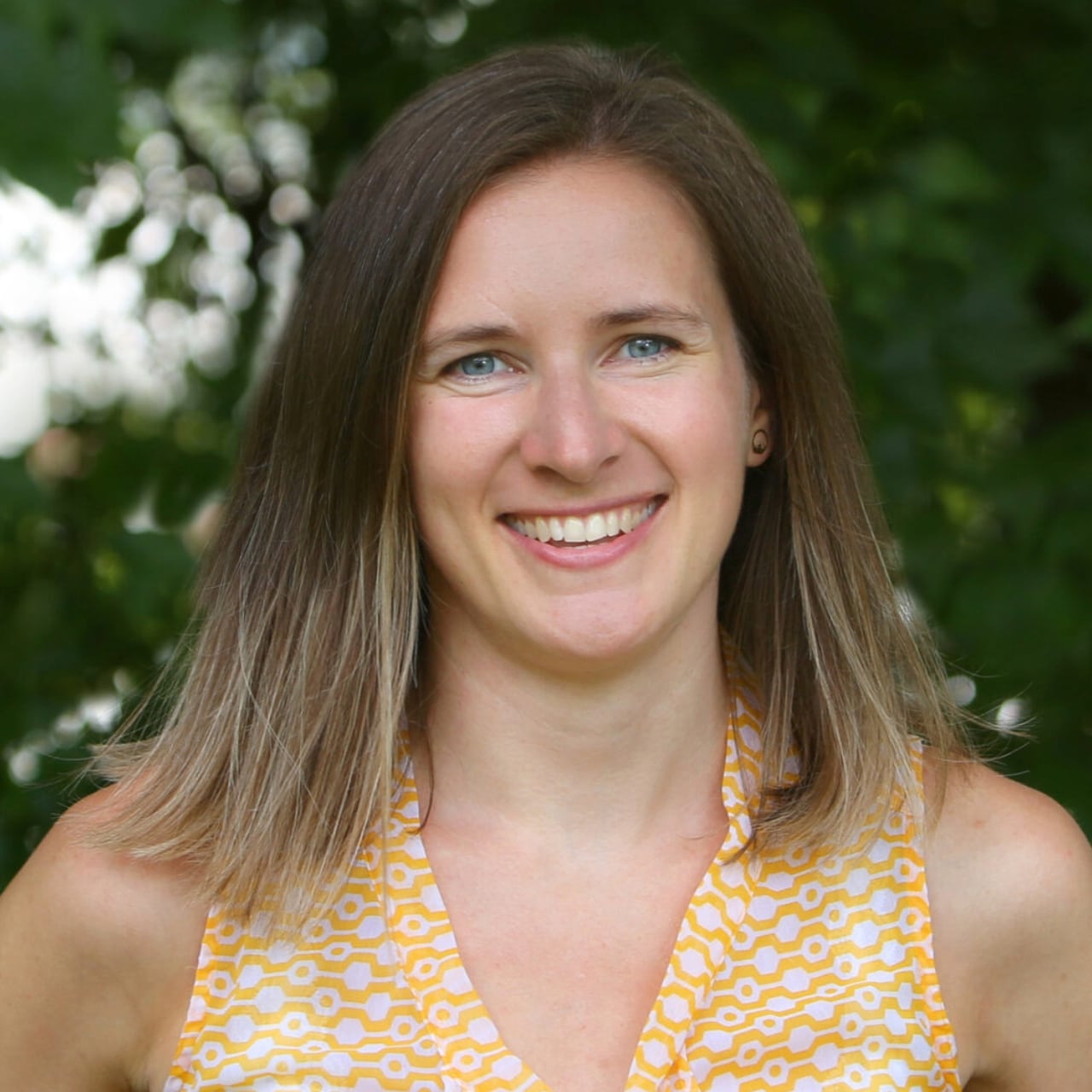 A woman with straight, shoulder-length brown hair and blue eyes smiles at the camera. She is wearing a sleeveless yellow patterned top and is standing outdoors in front of green leafy trees.