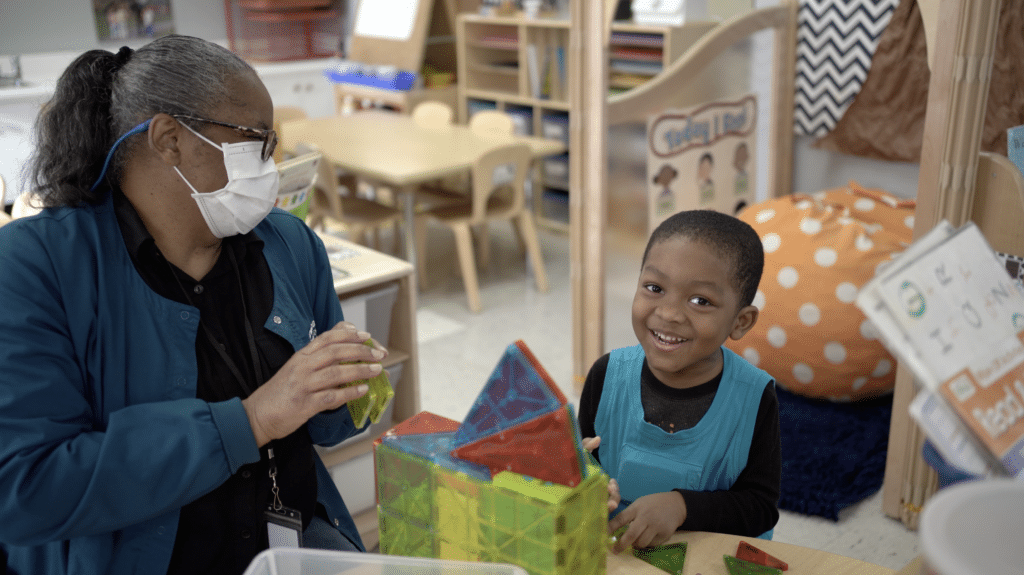 An adult wearing a mask and glasses plays with colorful magnetic tiles alongside a smiling young child in a classroom setting with tables, chairs, and educational materials.