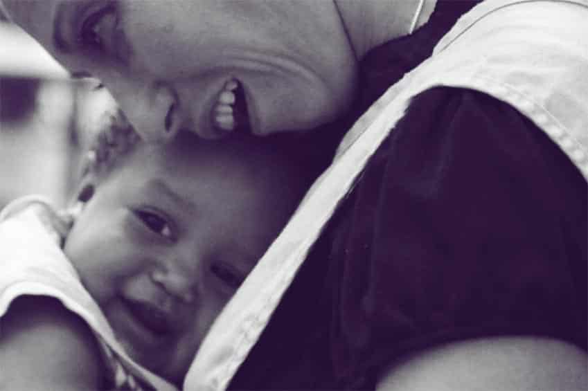 A woman lovingly hugs a smiling baby, who rests their head on her chest. Both are shown in close-up, capturing a tender, joyful moment. The image is in black and white.