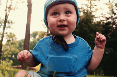 A smiling baby wearing a blue shirt and a blue hat with a chin strap sits on grass outdoors, surrounded by trees and sunlight filtering through the leaves.