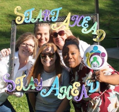 Five women smiling outdoors, holding a decorated frame that says "Start Ahead Stay Ahead." The background is grassy and sunny, and they appear joyful, enjoying a group moment together.