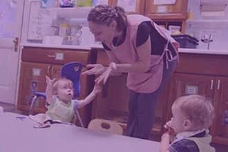 A caregiver in a pink apron bends down to interact with a toddler seated at a table while another toddler sits nearby in a childcare setting.