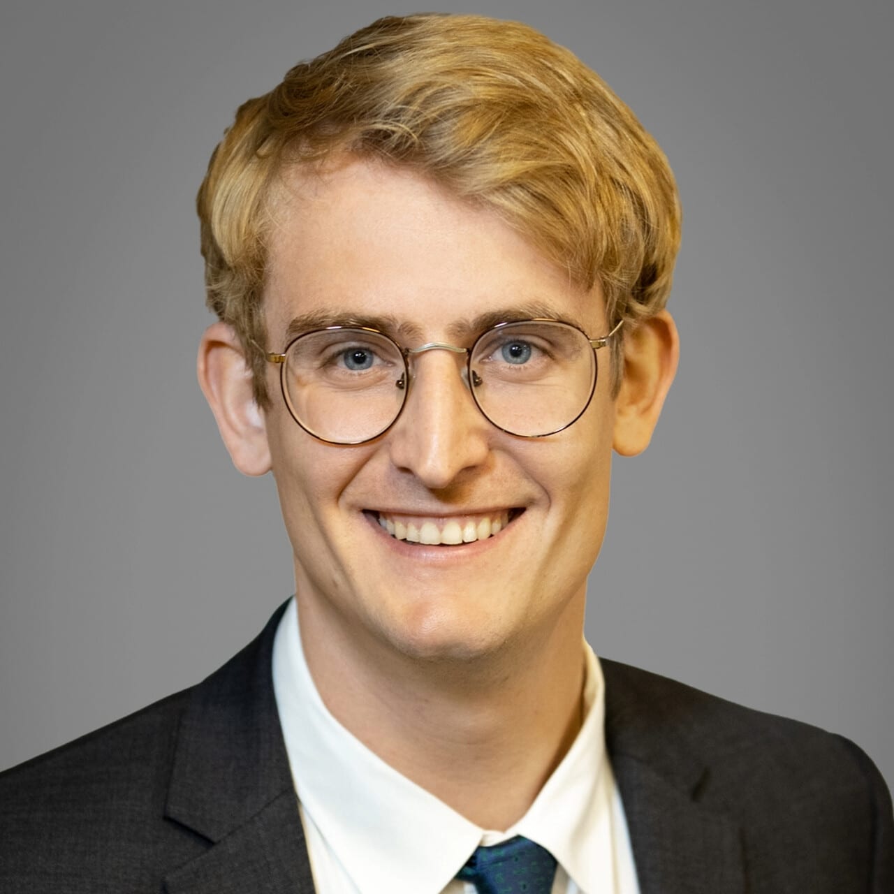 A young man with blond hair and round glasses, wearing a dark suit, white shirt, and blue tie, smiles at the camera against a plain gray background.