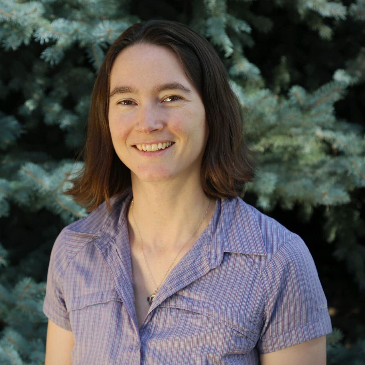 A woman with shoulder-length brown hair wearing a light purple plaid shirt smiles in front of green pine tree branches.