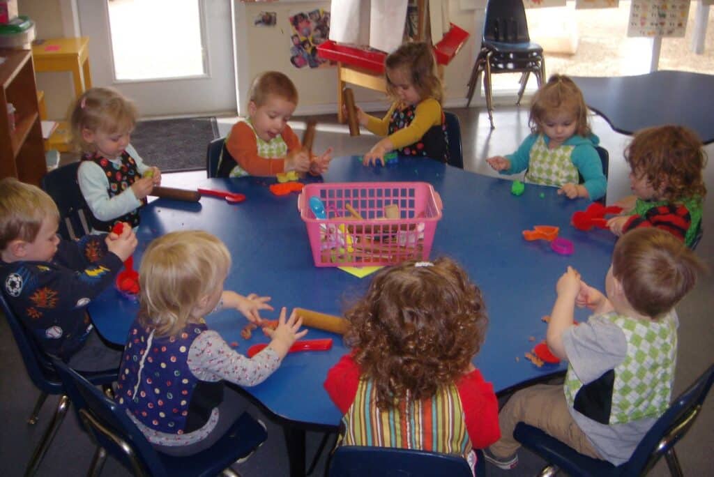 A group of young children sit around a blue table, playing with colorful playdough and tools. They are wearing aprons and focusing on their activities in a bright classroom.