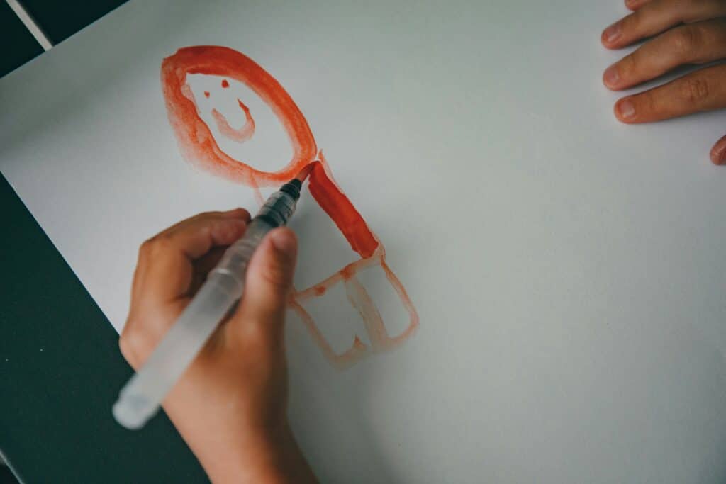 A child’s hand uses a water brush to paint a simple figure with red hair on white paper. Another hand rests on the edge of the paper. The drawing features a smiling face and a basic body outline.