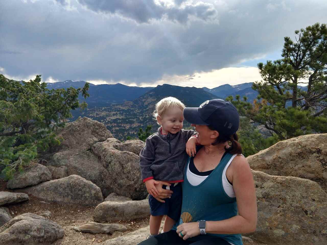 A woman and a young child smile at each other while sitting on rocky terrain with trees and mountains in the background under a cloudy sky.