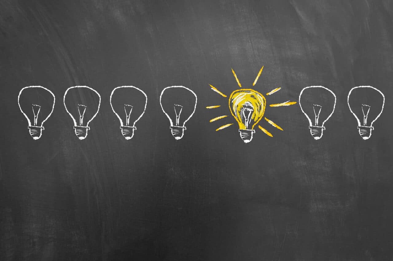 Six lightbulbs drawn in chalk on a blackboard; five are unlit, outlined in white, and the middle one is glowing brightly with yellow highlights, symbolizing a standout idea or innovation.