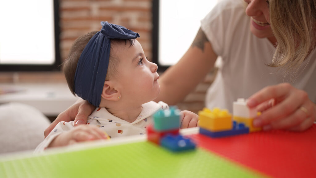 A baby wearing a blue headband looks up at an adult who is smiling and playing with colorful building blocks on a table. The scene is warm and indoors, with a blurred brick wall in the background.
