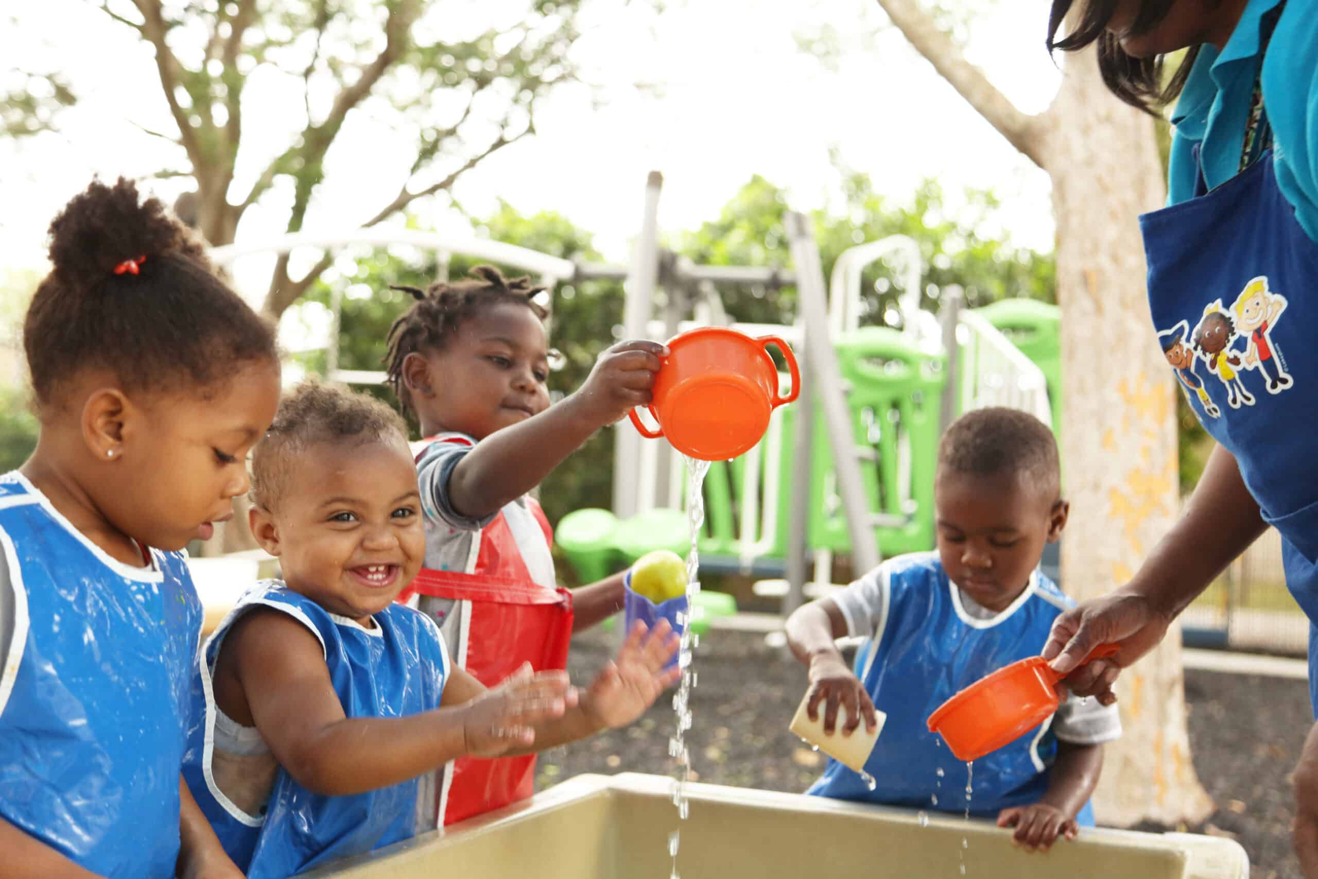 Four young children wearing blue and red aprons play with water and plastic cups outdoors, supervised by an adult. The children are smiling and laughing around a water table on a sunny day.