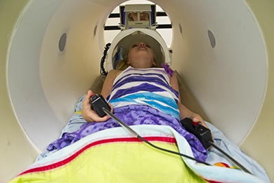 A young girl lies inside a medical imaging machine, possibly an MRI scanner, holding devices in both hands. She is covered with a colorful blanket and looking up, with her head positioned inside the scanner helmet.