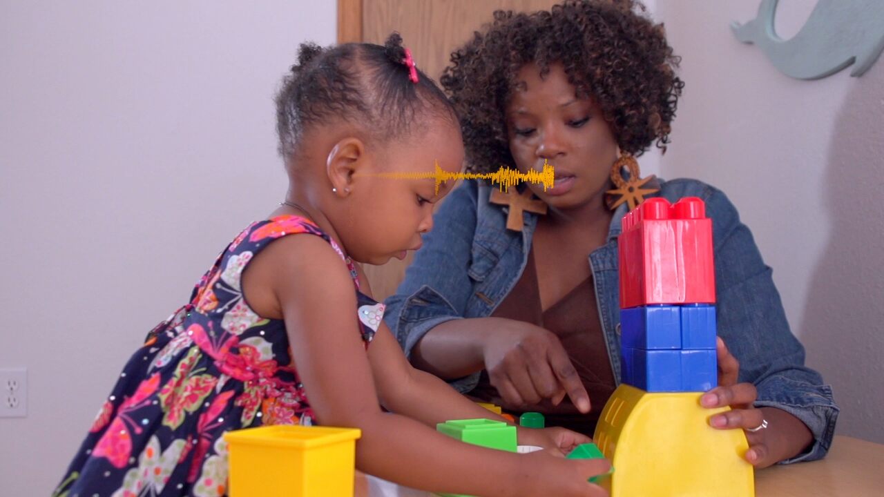 A woman and a young girl play with colorful plastic building blocks at a table. The woman points at the blocks while the girl, wearing a floral dress, focuses on stacking them.