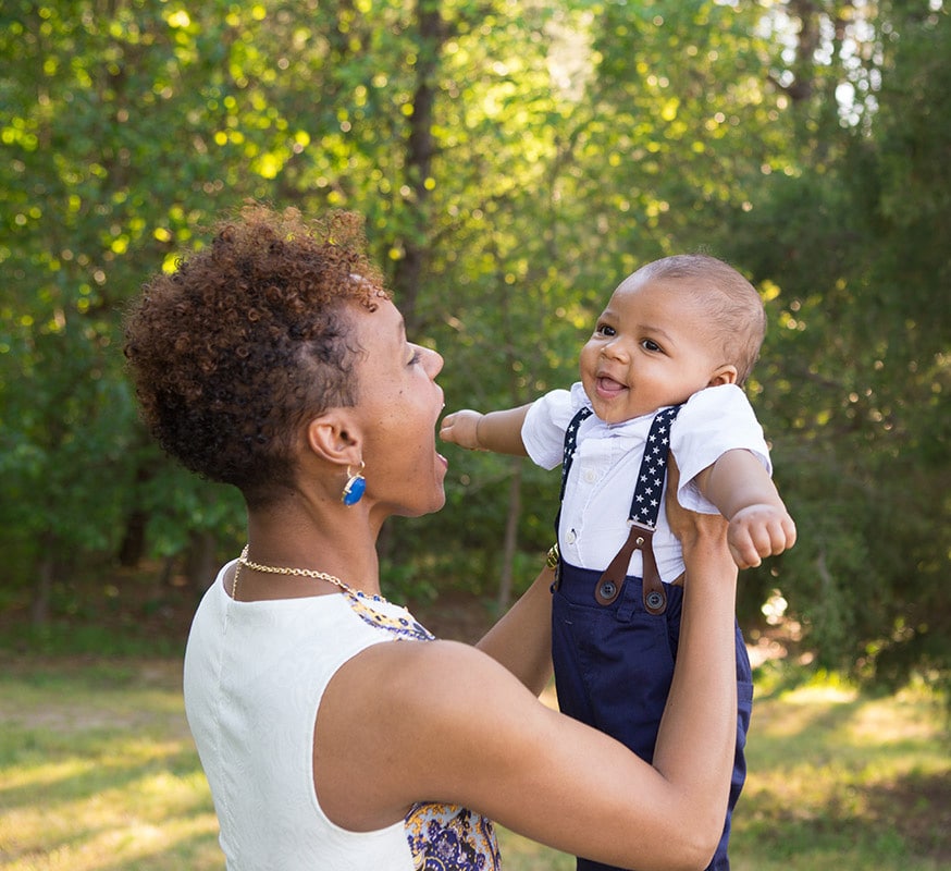 A woman smiles joyfully while lifting a laughing baby dressed in suspenders and a white shirt outdoors, surrounded by lush green trees.