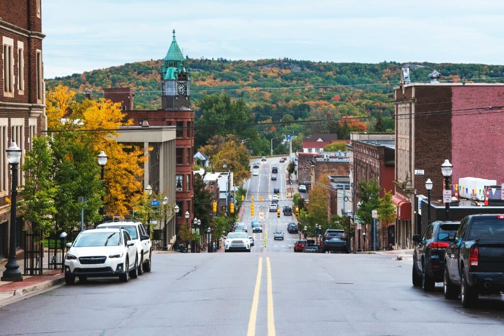 A small town street lined with parked cars and historic buildings, leading to a distant tree-covered hill with autumn foliage under a cloudy sky.
