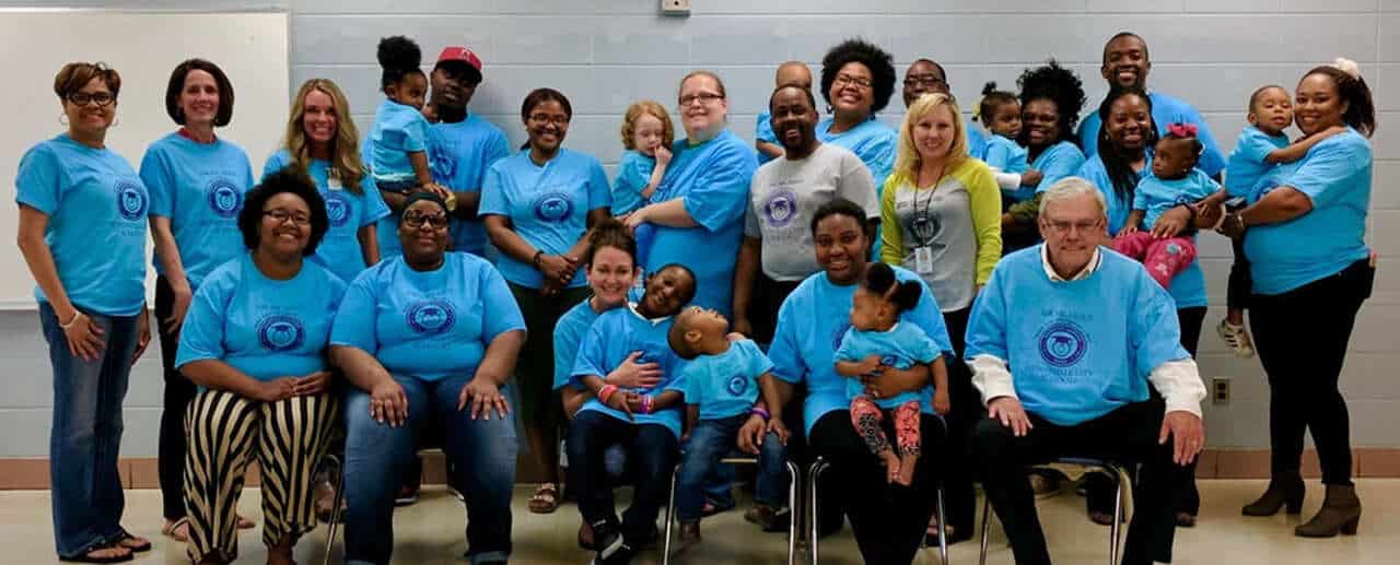 A diverse group of adults and children, many wearing matching blue T-shirts, pose together in a classroom setting. Some are seated while others stand behind them, smiling at the camera.