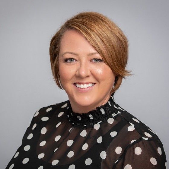 Smiling woman with short, light brown hair wearing a black sheer blouse with large white polka dots, posing against a plain light gray background.
