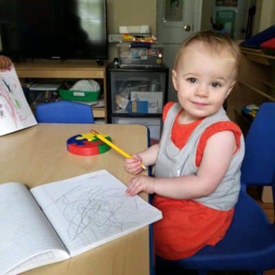 A young child in an orange dress sits at a table, holding a pencil and smiling. An open notebook with scribbles is in front of them, and colorful puzzle pieces are on the table.