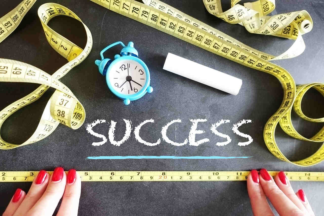 A black surface with the word "SUCCESS" written in chalk, surrounded by yellow measuring tapes, a blue alarm clock, a piece of chalk, and two hands with red nails holding a measuring tape at the bottom.