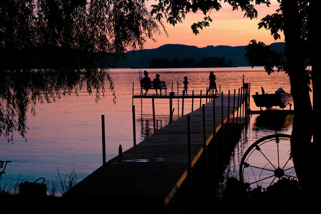 A wooden dock stretches into a calm lake at sunset, with silhouettes of people sitting and standing, trees framing the scene, and a boat moored to the dock. The sky is pink and orange, reflecting on the water.