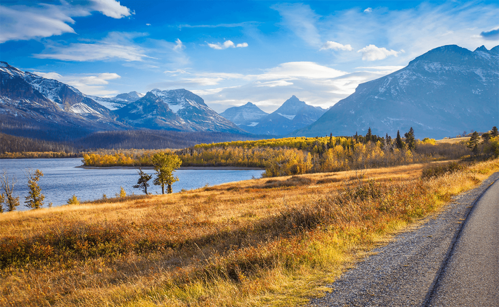 A scenic landscape featuring a clear blue lake, golden autumn trees, grassy fields, and distant snow-capped mountains beneath a partly cloudy sky. A paved road runs along the right side of the image.