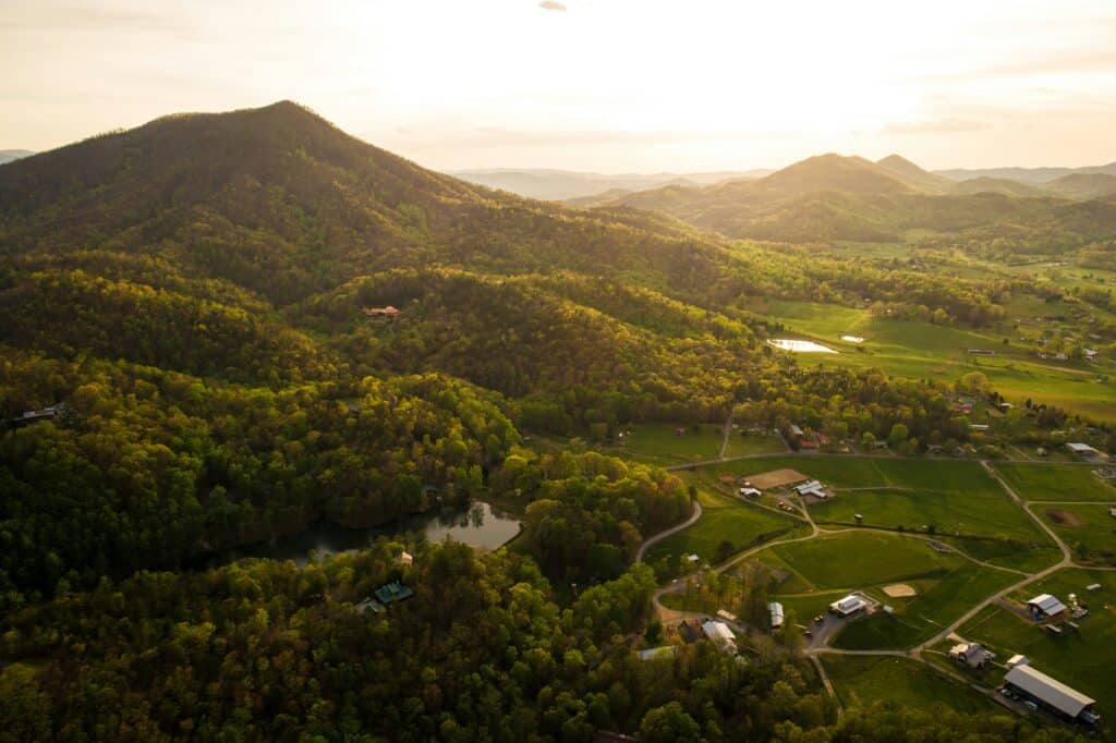 Aerial view of rolling green hills and valleys at sunset, with scattered houses, winding roads, small ponds, and tree-covered mountains in the background.