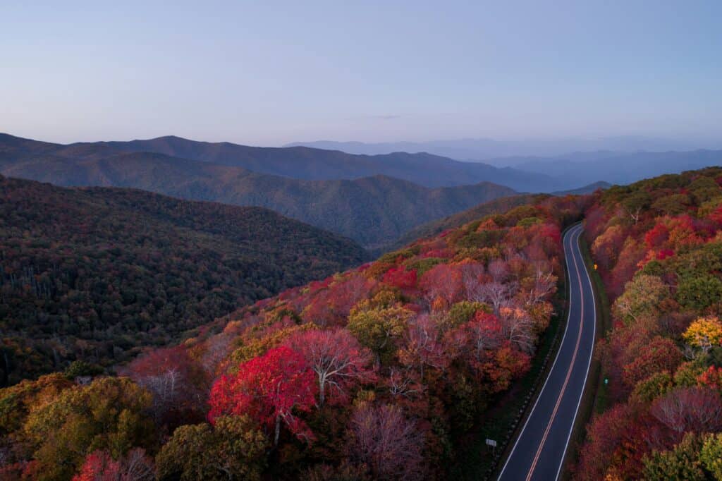 A winding road cuts through a forested mountain landscape with vibrant autumn foliage in shades of red, orange, and green under a clear sky. Rolling hills and distant blue mountains are seen in the background.