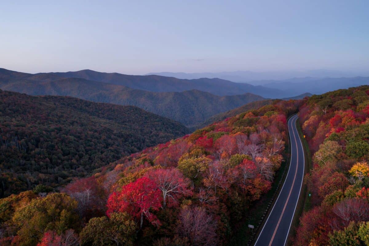 A winding road cuts through a forested mountain landscape with vibrant autumn foliage in shades of red, orange, and green under a clear sky. Rolling hills and distant blue mountains are seen in the background.