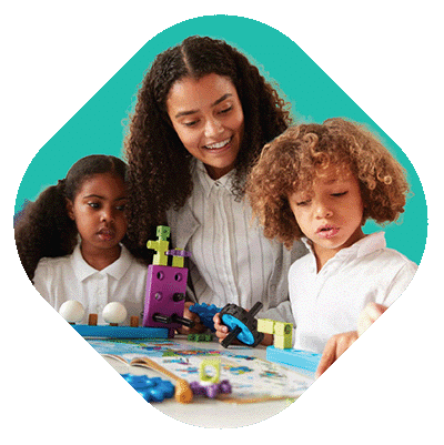 A woman helps two young children with curly hair build something with colorful educational toys at a table. All three are wearing white shirts and smiling, focused on the activity. The background is teal.