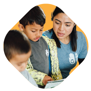 A woman and two young boys, all wearing shirts with hand-and-heart logos, look at a tablet together against an orange background.