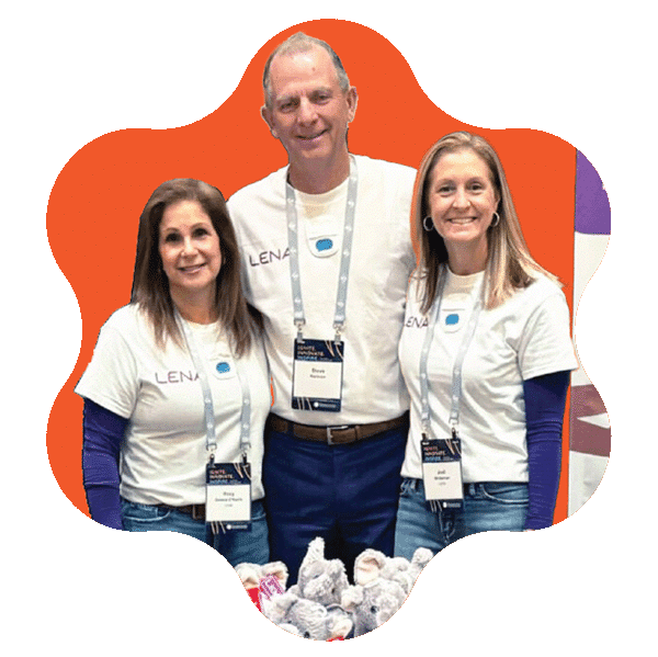Three people wearing white LENA t-shirts and conference badges stand together smiling in front of an orange background, with a table of stuffed animals in front of them.
