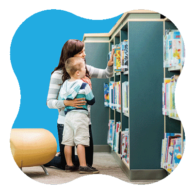 A woman and a young boy are kneeling in front of bookshelves, selecting a book together in a brightly lit library. Books are neatly arranged on the shelves.