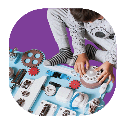A young child wearing grey pajamas plays with gears, switches, and a rotary dial on a busy board against a purple background.