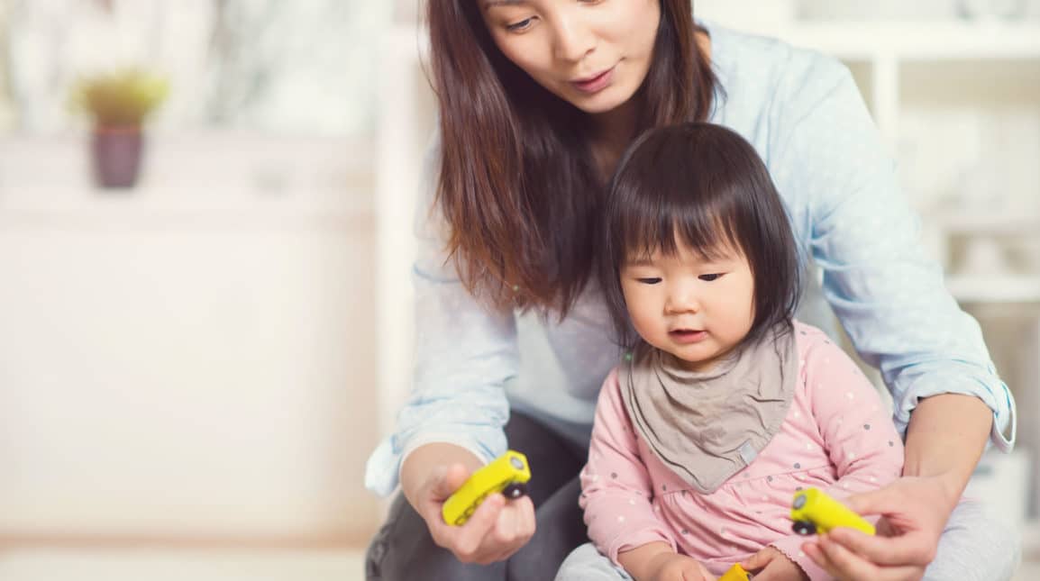 A woman and a young child sit together indoors, playing with small yellow toy cars. The woman is guiding the child, who is wearing a pink outfit and a gray bib. They both appear focused and engaged.