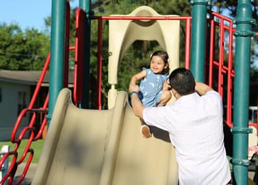 A young child in a blue dress slides down a playground slide while an adult, wearing a white shirt, waits at the bottom to catch her. Trees and a building are visible in the background.