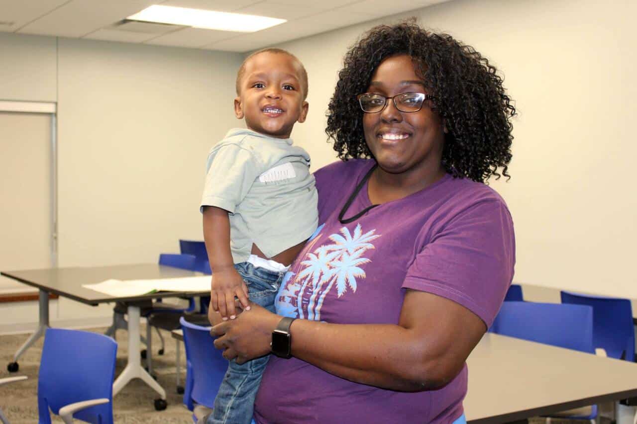 A smiling woman with curly hair, wearing glasses and a purple shirt, holds a happy young boy in a casual classroom setting with blue chairs and tables in the background.