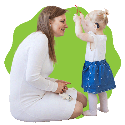 A woman kneels and smiles at a young girl with a hearing aid, who is standing and holding up a toy. They are interacting happily against a green background.