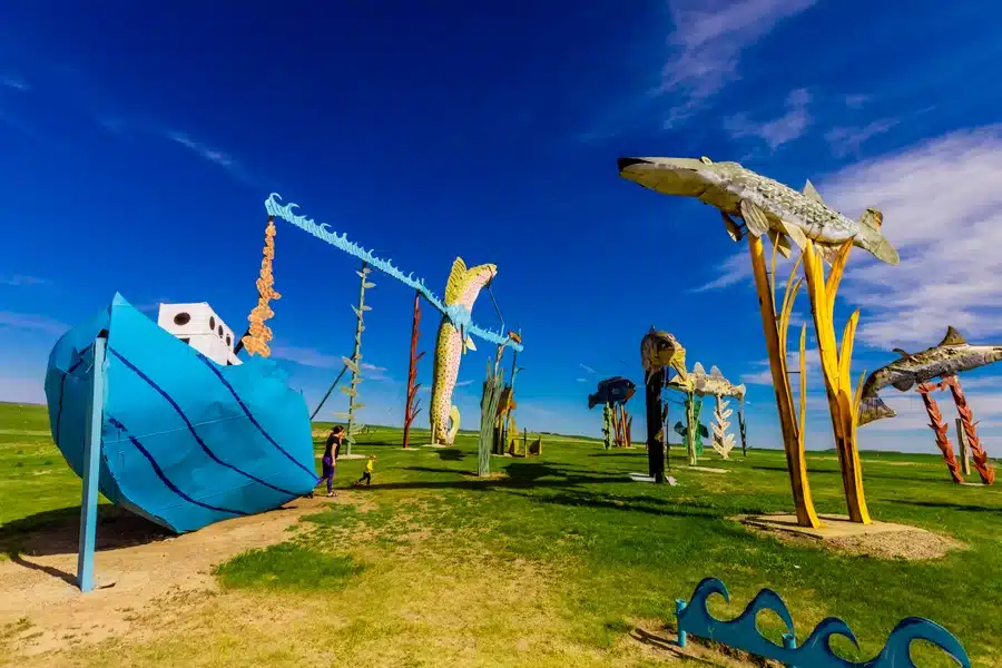 Colorful, whimsical metal sculptures of a blue boat and large fish stand on grassy land under a clear blue sky, with a few people walking nearby and vibrant yellow and blue accents throughout the artwork.