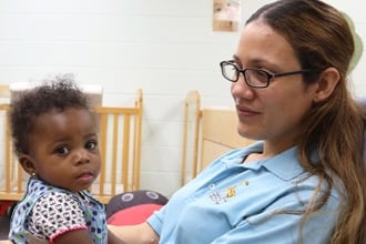 A woman with glasses, wearing a light blue shirt, holds a young child with curly hair and a patterned outfit, seated in a room with a crib and light-colored walls in the background.
