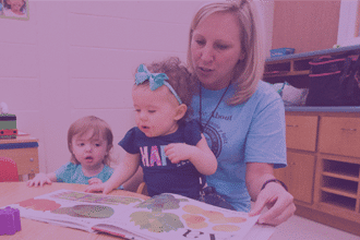 An adult woman sits with two young children in a classroom, reading a colorful picture book together. One child wears a blue bow headband and the other has a blue shirt and brown hair.
