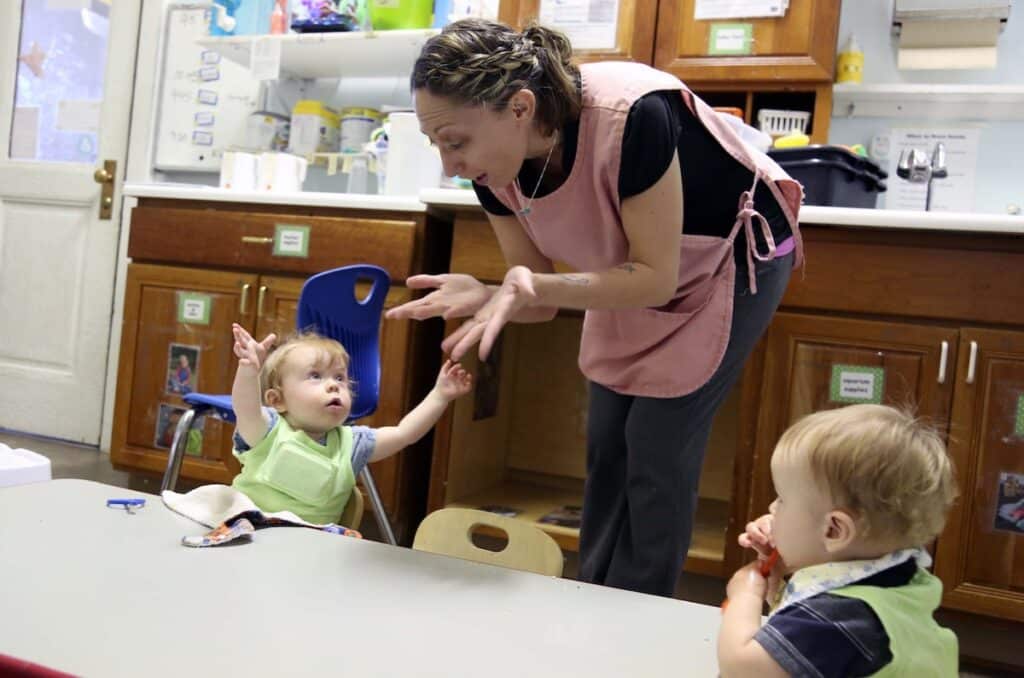 A caregiver in a pink apron interacts with a toddler raising their arms while another toddler sits nearby at a table in a daycare classroom with wooden cabinets and children’s artwork on the walls.
