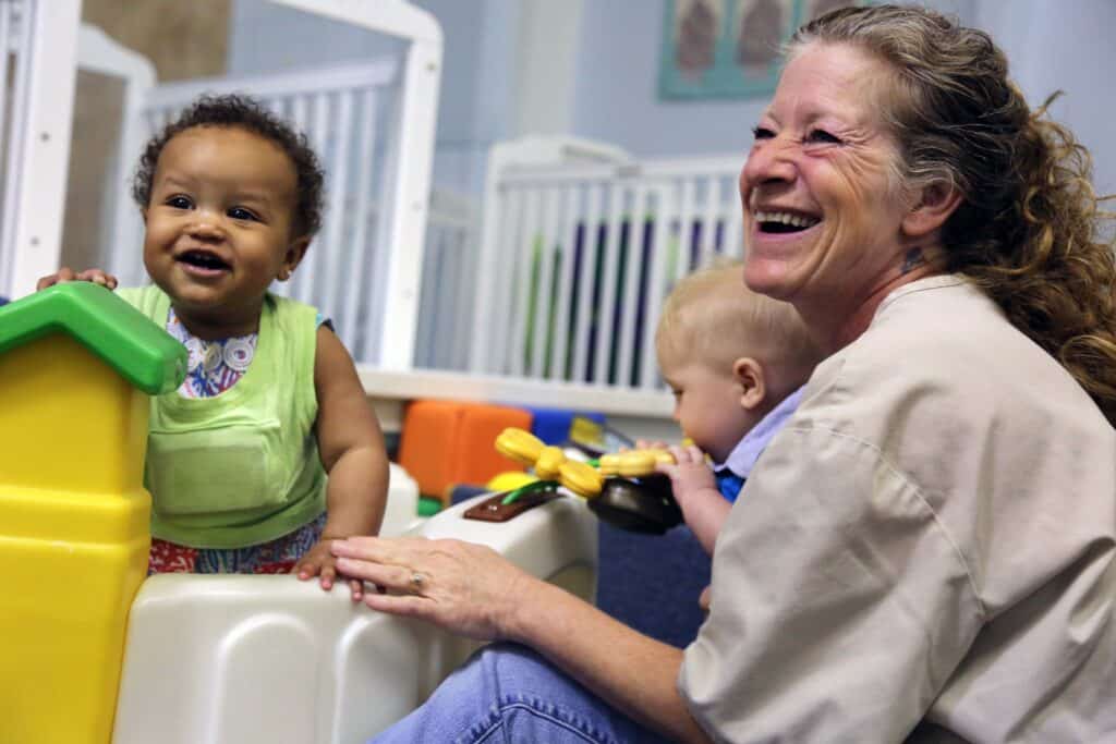 A smiling woman sits beside two infants in a play area. One baby stands by a plastic play structure, smiling, while the other sits nearby holding a toy. The scene is cheerful and playful.