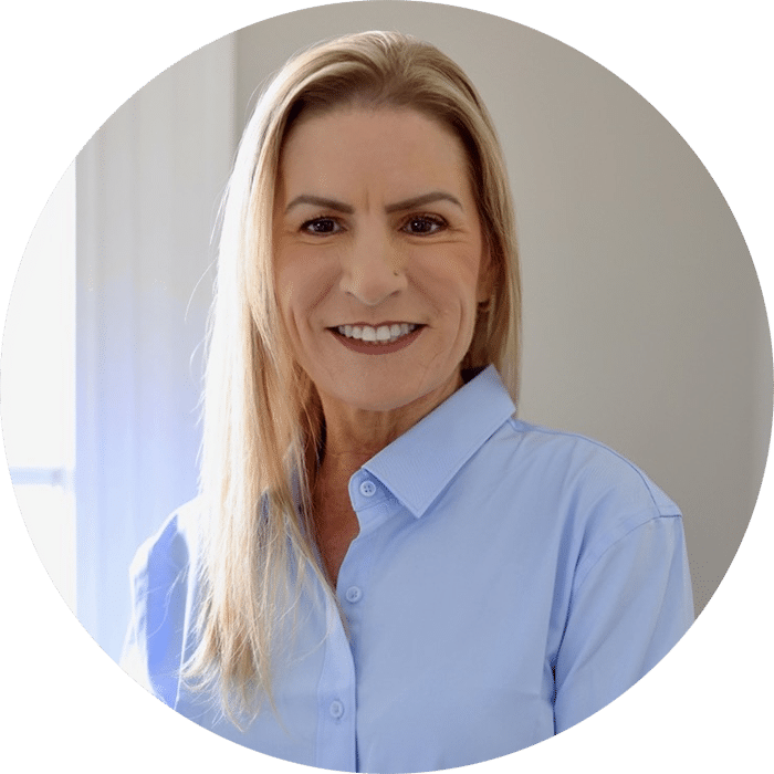 A woman with long blonde hair wearing a light blue button-up shirt smiles while standing indoors near a sunlit window.