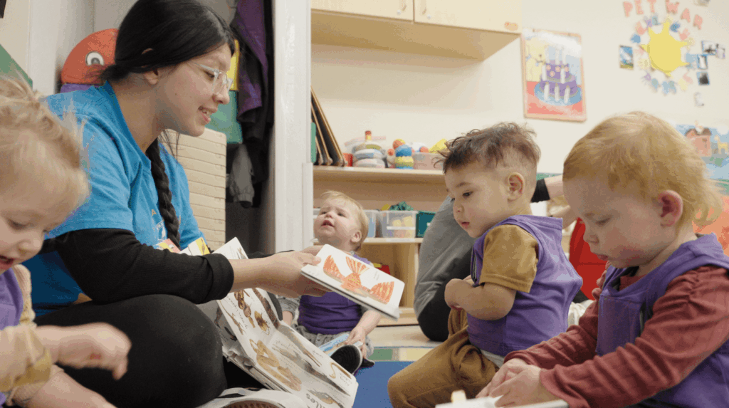 A caregiver reads a picture book to a group of toddlers sitting on the floor in a colorful classroom. The children are engaged and wearing purple bibs, surrounded by toys and books.
