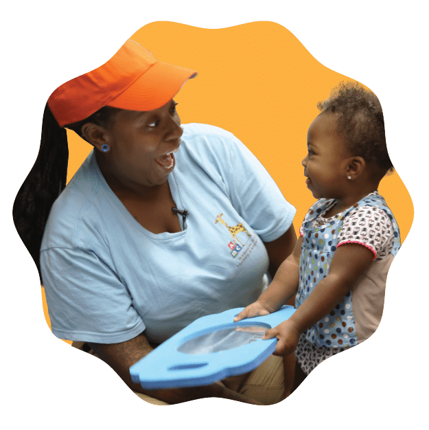 A smiling woman in an orange cap and blue shirt joyfully interacts with a smiling toddler, who is holding a blue toy, against an orange background.