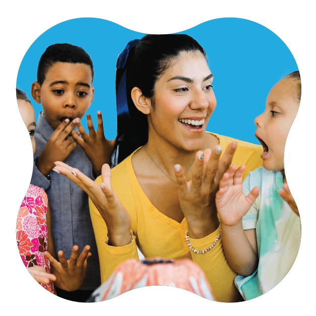 A woman and three children excitedly gather around a table, smiling and interacting with each other, against a bright blue background.